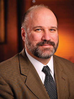 Andrew Houtenville is a man with very short gray hair and a beard wearing a brown suit