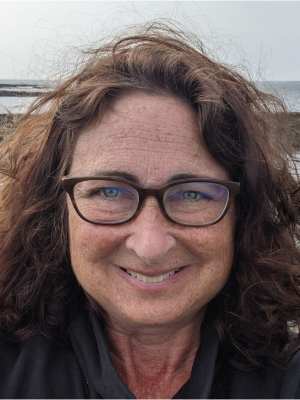 Deb Brucker is a woman with auburn hair and glasses pictured in front of the ocean