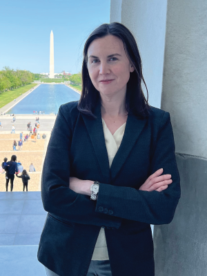 Megan Henly is a woman with brown hair in a suit jacket standing with the Washington Monument and reflection pool in the background