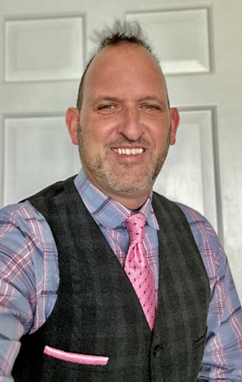 Andrew Karhan is a man with short brown hair and beard, wearing a tweed vest over a bright flannel with a vibrant pink patterned tie