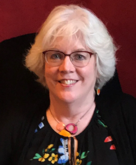 Leslie Jones is a woman with short, white hair wearing red rimmed glasses and a colorful necklace and blouse.