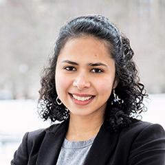Monica Farid is a woman with dark curly hair and black jacket, smiling a the camera.