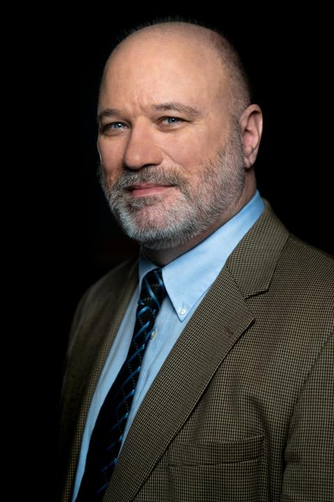 Andrew Houtenville is a man with very short hair and a salt and pepper beard wearing a tweed suit jacket and light blue shirt with a dark blue striped tie