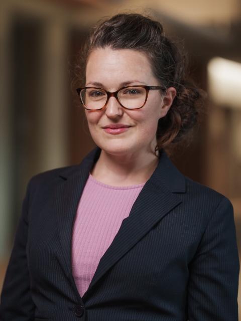 Stacia Bach is a professionally dressed woman with brown wavy hair pulled back and wearing wide rimmed glasses.